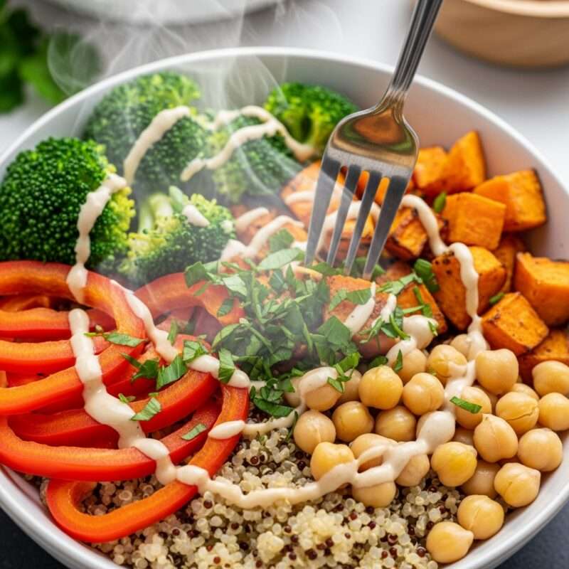 
Buddha bowl with steaming broccoli, roasted sweet potato, red bell peppers, chickpeas, quinoa, and tahini drizzle with a fork.